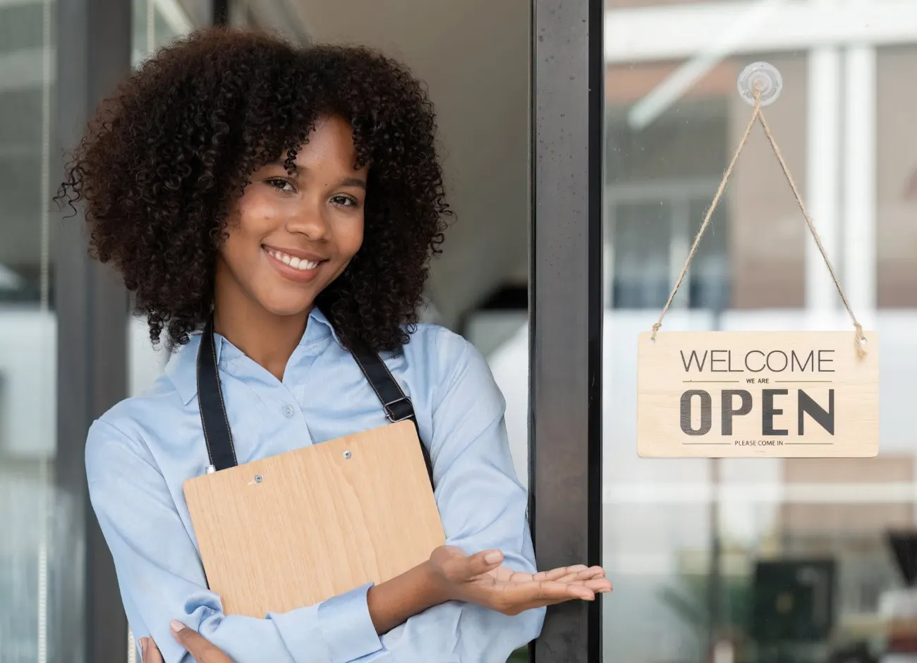 Smiling small business owner in a denim apron holding a clipboard at the door of her café, gesturing to a Welcome, we are open sign
